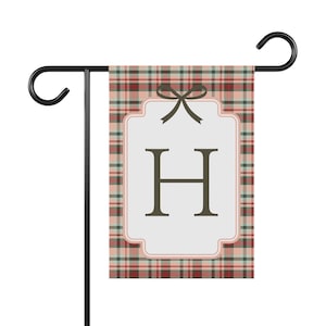 Könnte beinhalten: Eine Gartenflagge mit einem rot, grün und cremefarbenen Karomuster. Sie hat ein weißes Etikett mit dem dunkelbraunen Buchstaben "H" und einer Schleife. Die Flagge wird auf einem schwarzen Metallständer präsentiert.