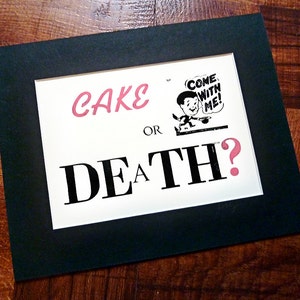 Peut inclure: Une impression encadrée noire avec un fond blanc et le texte "CAKE OR DEATH?" en noir avec un point d'interrogation rose. Une petite illustration en noir et blanc d'un homme avec le texte "COME WITH ME!" est dans le coin supérieur droit.