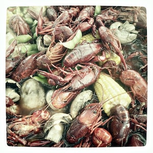 May include: A close-up of a seafood boil with cooked crawfish, sweetcorn, green vegetables, and mushrooms. The crawfish are a deep red colour, and the sweetcorn is bright yellow. A lime wedge is visible.