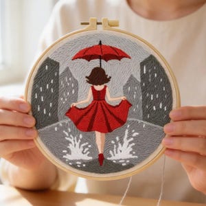 Peut inclure: Broderie sur un cercle de bois représentant une femme en robe rouge sous un parapluie rouge, marchant sous la pluie. Des bâtiments gris et des éclaboussures blanches complètent la scène. Le mot "AURELI" est inscrit en haut.