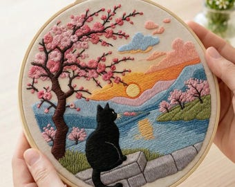 May include: Embroidered hoop art featuring a black cat overlooking a serene landscape with a cherry blossom tree, a sunset, and a body of water. The word "AURELI" is at the top. The artwork is held by two hands.