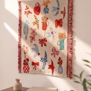 Puede incluir: Un tapiz rectangular tejido con fondo beige y motivos bordados en rojo, azul y naranja que incluyen pájaros, flores, jarrones y lazos. El tejido presenta un borde con flecos en hilos rojos y blancos y el texto AURELI.