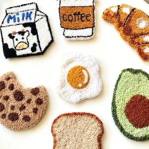 Puede incluir: Colección de parches de tela hechos a mano con forma de alimentos y bebidas para el desayuno. Incluye una caja de leche, una taza de café, un cruasán, una galleta, un huevo frito, un aguacate y una rebanada de pan tostado. La palabra "AURELI" está impresa.