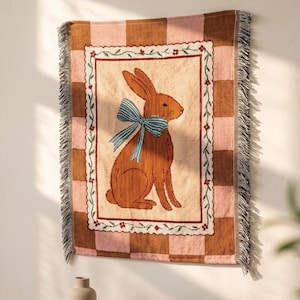 May include: A woven wall tapestry featuring a brown rabbit with a blue striped bow around its neck, centred within a floral border and a brown and pink checkered background. The word AURELI is printed in white at the top of the textile piece.