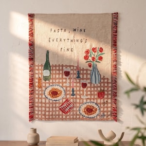 Könnte beinhalten: Ein rechteckiger, gewebter Wandbehang mit beigem Hintergrund und braunem Karomuster. Der Text lautet PASTA, WINE EVERYTHING'S FINE. Er zeigt Pastateller, Weingläser, eine Flasche und eine Vase mit roten Blumen auf einem Tisch.