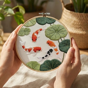 May include: Embroidered hoop art featuring koi fish and lily pads. The design includes orange, white, and black koi fish swimming among green lily pads. The embroidery is set in a wooden hoop, creating a framed artwork.
