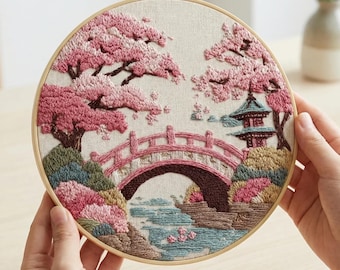Op de afbeelding: Geborduurd kunstwerk in een houten ring met een Japanse landschap met een roze brug, kersenbloesem bomen en een pagode. Het kunstwerk wordt door handen vastgehouden en heeft het woord "AURELI" bovenaan.