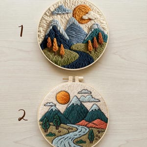 May include: Two round embroidery hoops with landscape scenes. One features mountains, a river, trees, and a sun. The other has a similar design with a river, mountains, trees, and a sun. The embroidery is done with various shades of blue, green, orange, and beige.