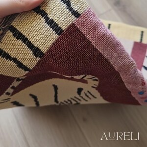 May include: Close-up of a woven textile with a geometric pattern in beige, burgundy, and black. The textile is folded, revealing a portion of a tiger design. The word "AURELI" is visible in the bottom right corner.
