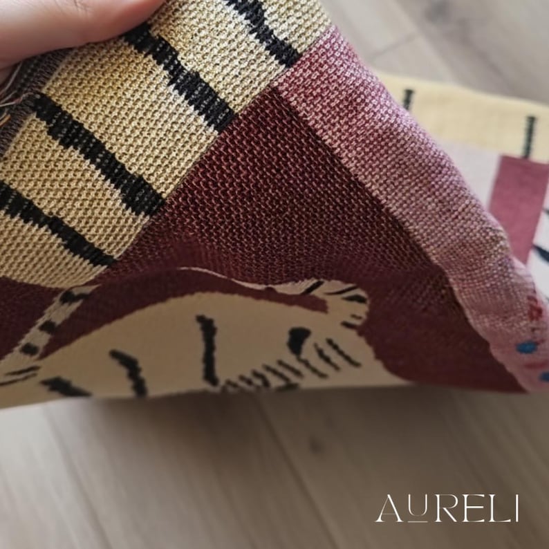 May include: Close-up of a woven textile with a geometric pattern in beige, burgundy, and black. The textile is folded, revealing a portion of a tiger design. The word "AURELI" is visible in the bottom right corner.