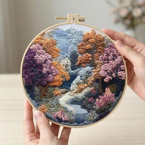 Puede incluir: Un bordado detallado de una escena de bosque en un aro de madera redondo. La obra de arte presenta un río que serpentea a través de árboles con follaje púrpura, naranja y rosa. La palabra "AURELI" está bordada en la parte inferior. El aro es sostenido por manos.