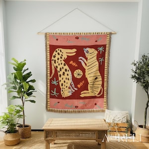 May include: A tapestry featuring two stylized big cats, a leopard and a tiger, against a red background. The tapestry is rectangular with a fringed border and is suspended from a wooden rod. The design includes fish bones and palm trees.