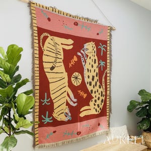 May include: A woven wall hanging featuring a tiger and a leopard in a stylized design. The tapestry has a red background with yellow and black animal figures, a pink border, and fringed edges. It is mounted on a wooden rod.