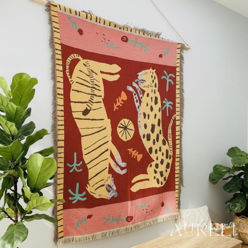 May include: A woven wall hanging featuring a tiger and a leopard in a stylized design. The tapestry has a red background with yellow and black animal figures, a pink border, and fringed edges. It is mounted on a wooden rod.