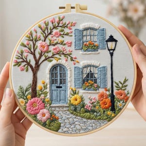 May include: Embroidered hoop art featuring a quaint cottage scene. The artwork showcases a white house with blue shutters and door, a cobblestone path, and vibrant flowers. A tree with pink blossoms and a black lamppost complete the charming design. The word "AURELI" is at the top.