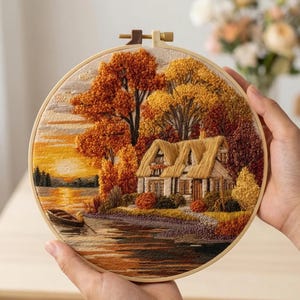 Op de afbeelding: Ronde borduurring met een gedetailleerd herfstlandschap. De scène toont een huis met een rieten dak, bomen met oranje en gele bladeren, een meer en een zonsondergang. Het woord "AURELI" staat bovenaan.