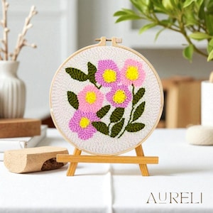 Op de afbeelding: Een bloemenborduurring met roze en gele bloemen en groene bladeren. De borduurring wordt gepresenteerd op een kleine houten ezel. Het woord "AURELI" is zichtbaar aan de onderkant, tegen een witte achtergrond.
