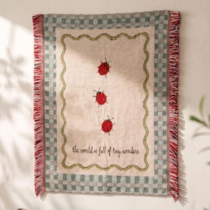 May include: A rectangular woven wall hanging featuring a cream-coloured centre with three red ladybugs and the text "the world is full of tiny wonders" in black script. The border is a green and cream checkerboard pattern with red fringe edges.
