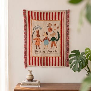 Peut inclure: Une tenture murale rectangulaire tissée avec des rayures verticales rouges et crème, présentant un panneau central brodé montrant un lapin, un chien et une souris se tenant la main. Le texte "Best of friends" est brodé sous les personnages.