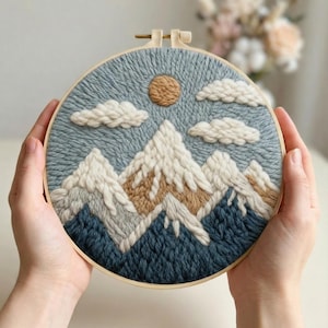 May include: A hand-stitched embroidery hoop art piece featuring a mountain landscape. The design includes snow-capped mountains in white and tan, a tan sun, and fluffy white clouds against a blue sky. The embroidery is set in a wooden hoop.