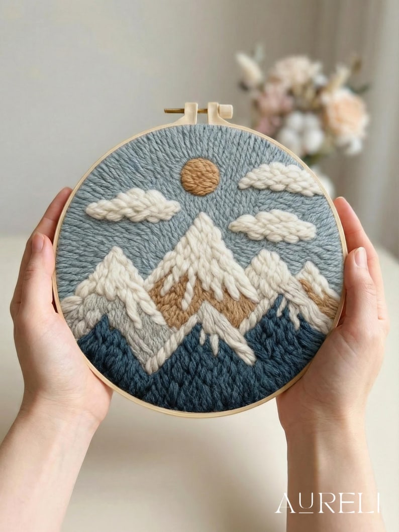 May include: A hand-stitched embroidery hoop art piece featuring a mountain landscape. The design includes snow-capped mountains in white and tan, a tan sun, and fluffy white clouds against a blue sky. The embroidery is set in a wooden hoop.