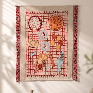 May include: A rectangular woven wall hanging featuring a red grid pattern on a beige background with the text AURELI at the top. The design depicts a picnic scene with a pizza, a pie, a charcuterie board, a baguette, fruit, and a bouquet of flowers.