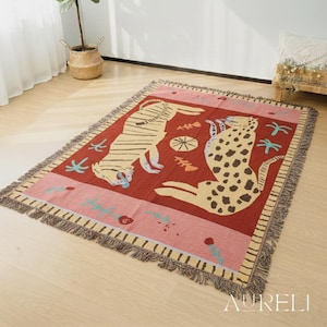 May include: A rectangular woven throw blanket featuring a tiger and a leopard design. The blanket has a red center with a pink border, fringed edges, and decorative elements in blue and beige. The word "AURELI" is in the bottom right corner.