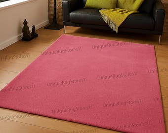 May include: A large, rectangular, solid pink rug. The rug is placed on a wooden floor, with a black leather sofa and green throw pillows in the background. The rug is a soft, plush material.