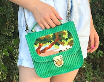 May include: A green crossbody bag with a woven texture and a gold clasp. The bag has a rectangular flap with a cross-stitch design of a tree and a landscape.