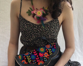 May include: A black floral embroidered handbag with a scalloped edge and a gold clasp. The bag is held by a person wearing a black and white floral dress and a colorful floral necklace.