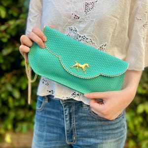 May include: A turquoise clutch with a gold chain strap and a gold horse emblem. The clutch has a scalloped edge with gold trim. The bag is made of a woven material.