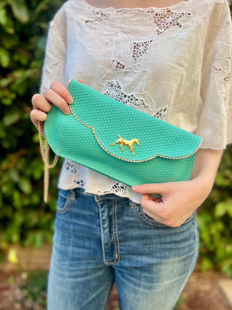 May include: A turquoise clutch with a gold chain strap and a gold horse emblem. The clutch has a scalloped edge with gold trim. The bag is made of a woven material.