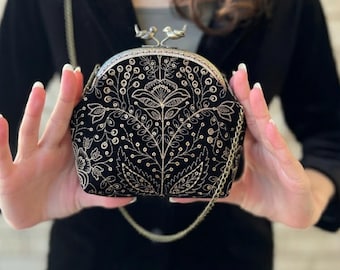 May include: Black velvet clutch with gold floral embroidery. The clasp features two small bird figures. A gold chain strap is attached. The bag is held by a person.