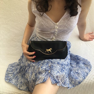 May include: Black satin clutch with a gold-colored horse emblem. The clutch has a scalloped edge and a curved design. The horse emblem is centered on the front of the clutch. The clutch is a classic accessory.