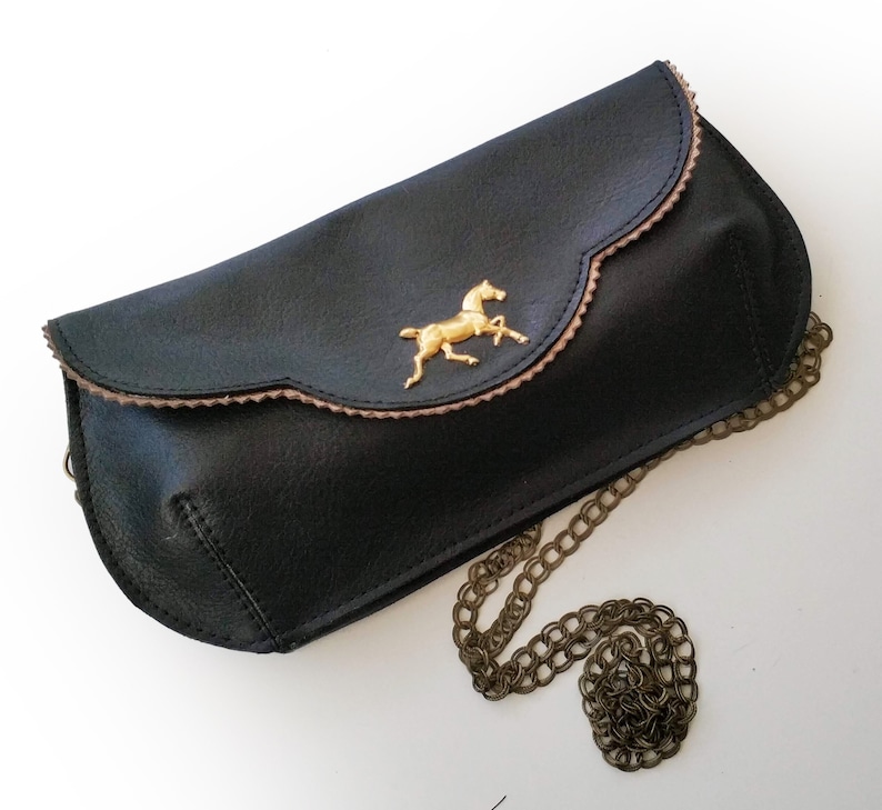 May include: Black clutch purse with a gold-colored horse emblem. The purse has a scalloped edge with a brown trim and a chain strap. The purse is made of a textured material.