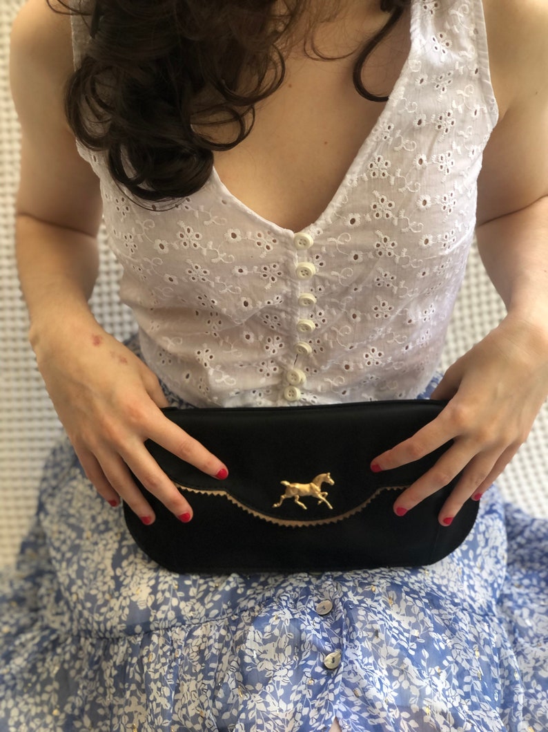May include: Black satin clutch with a gold-colored horse emblem. The clutch has a scalloped edge with gold trim. The background includes a white eyelet top and a blue floral skirt.