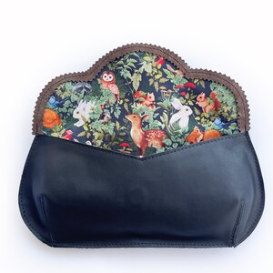 May include: A black leather clutch with a woodland-themed fabric lining. The lining features a pattern of forest animals, including deer, rabbits, and squirrels, set against a backdrop of lush greenery. The clutch has a scalloped edge.