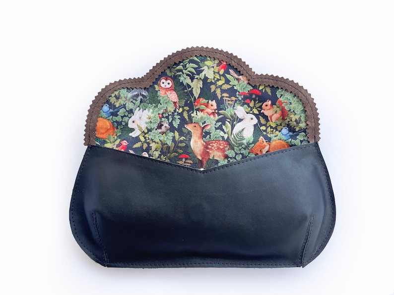 May include: A black leather clutch with a woodland-themed fabric lining. The lining features a pattern of forest animals, including deer, rabbits, and squirrels, set against a backdrop of lush greenery. The clutch has a scalloped edge.