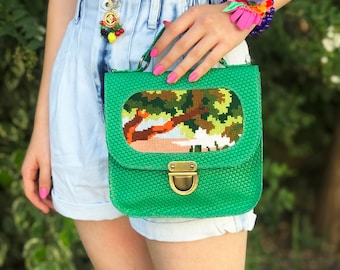 May include: A green woven crossbody bag with a floral embroidered patch and a gold clasp. The bag has a woven texture and a rectangular shape.