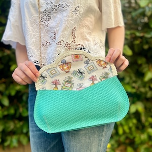 May include: A turquoise clutch with a gold chain strap. The interior lining features a perfume bottle pattern in various colors. The clutch is held open, revealing the patterned lining. The exterior has a textured appearance.