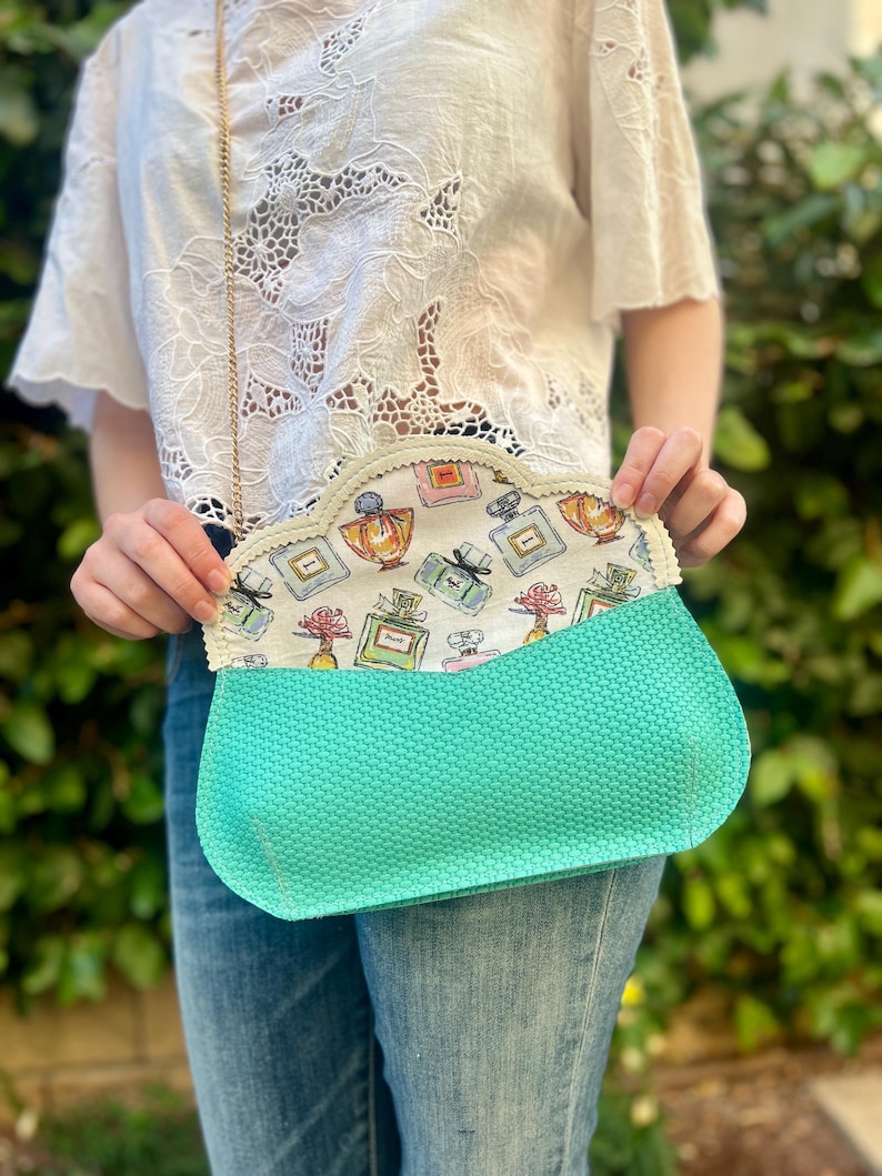 May include: A turquoise clutch with a gold chain strap. The interior lining features a perfume bottle pattern in various colors. The clutch is held open, revealing the patterned lining. The exterior has a textured appearance.