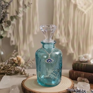 May include: A turquoise glass bottle with a diamond-shaped stopper and a silver chain with an evil eye pendant. The bottle sits on a wooden slice, with dried flowers and crystals in the background. The bottle has a textured leaf design.
