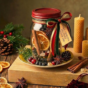 Può includere: Un barattolo di vetro pieno di fette d'arancia essiccate, bastoncini di cannella, anice stellato e bacche, sormontato da un coperchio rosso e un nastro decorativo. Un'etichetta recita "Yule | Winter Solstice | Summer Jar". Il barattolo è su una superficie di legno con candele.