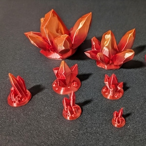 May include: A collection of red and orange crystal-shaped decorative objects. The crystals vary in size, with some larger, flower-like formations and smaller, individual pieces. The objects are arranged on a dark surface.