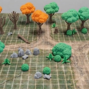 Op de afbeelding: Een tabletop gaming terreinset met miniatuurbomen met oranje en groen blad, bruine stammen, grijze rotsen en kleine groene struiken, gerangschikt op een bedrukte rastermat met een zandpadontwerp voor rollenspellen.