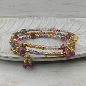 May include: A multi-strand wrap bracelet featuring a mix of gold-tone metallic beads, translucent purple, pink, and yellow glass beads, and faceted crystal accents. The design includes two dangling charms at the ends, each with a small bead.