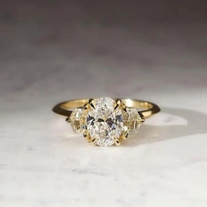 May include: A gold engagement ring with a large oval-cut diamond center stone and two smaller side stones. The ring has a classic, elegant design, perfect for a special occasion. The diamonds sparkle brightly against the gold band.