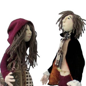 May include: Two handmade dolls, one wearing a burgundy hooded dress with a patterned skirt and the other wearing a black jacket with a burgundy scarf and trousers.