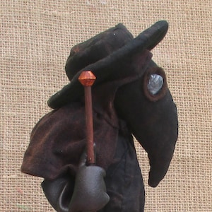 Peut inclure: Une poupée en feutre noir portant un long manteau noir et un chapeau noir avec un long bec. La poupée tient un bâton en bois avec un dessus brun et orange.