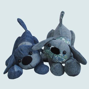 May include: Two plush dog toys crafted from blue denim fabric. The left dog is solid dark blue denim with black button eyes and a black nose. The right dog features light blue denim with a floral pattern on the face and neck, black button eyes, and a black nose.